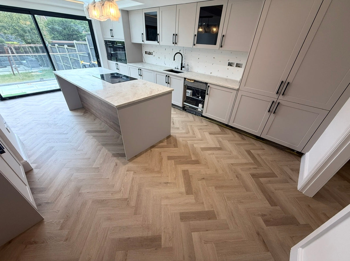 A modern kitchen interior with white cabinetry, a light marble-look island countertop, and light oak herringbone wood flooring. Large glass doors on the left reveal a backyard patio and greenery.