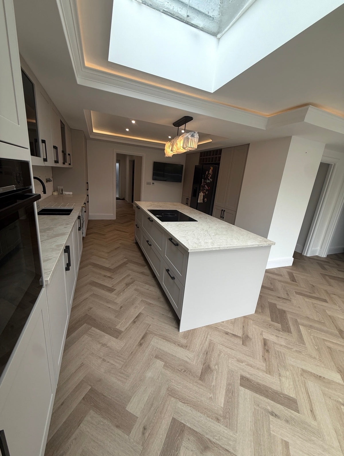A modern kitchen interior with white cabinetry, a white island with a light stone countertop, herringbone wood flooring, recessed lighting, and a large skylight.