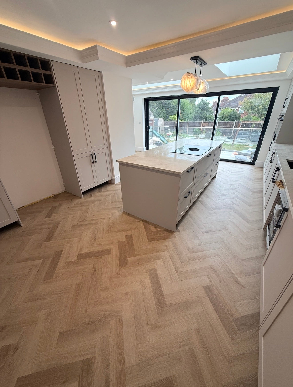 A modern kitchen interior with light oak herringbone wood flooring, white cabinetry, and a central island, photographed from an elevated floor-to-ceiling window view.