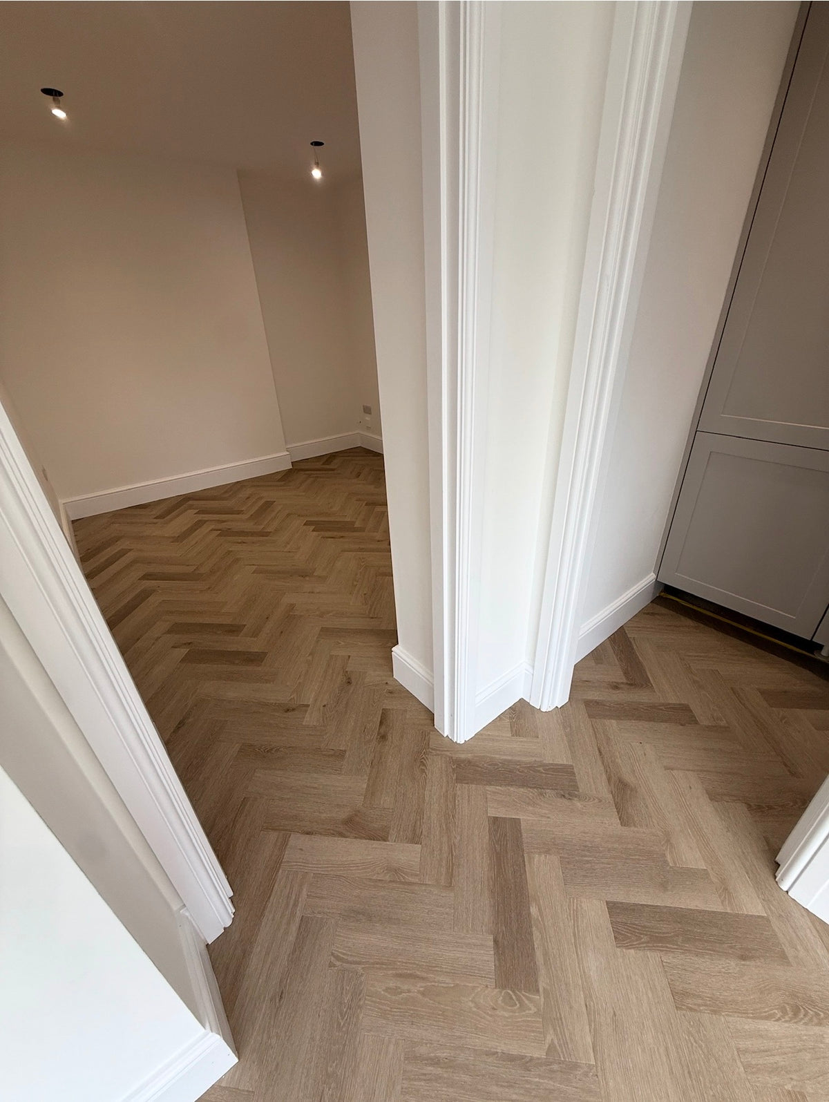 A herringbone-patterned light oak wood floor in a modern room with white walls, white baseboards, and white door frames.