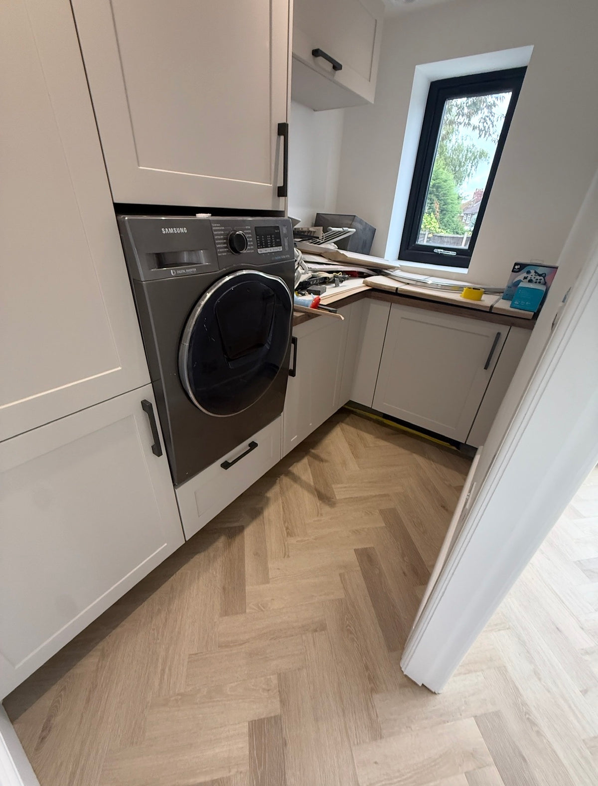A Samsung washing machine in a dark gray finish installed in a modern white built-in wardrobe cabinet, set against a light wood herringbone floor.