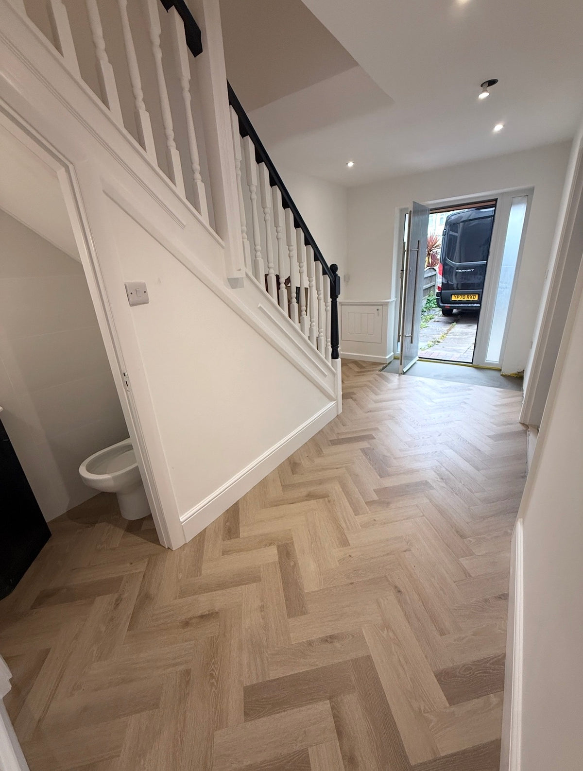 A bright, modern interior hallway featuring light oak wood herringbone parquet flooring, white walls, a white staircase with dark wooden railings, and an open doorway in the background.