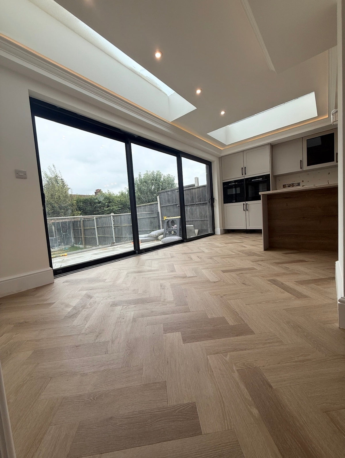 A modern interior room with light oak herringbone hardwood flooring, featuring a large black-framed sliding glass door, white kitchen cabinets, and a wood-look island in the background.
