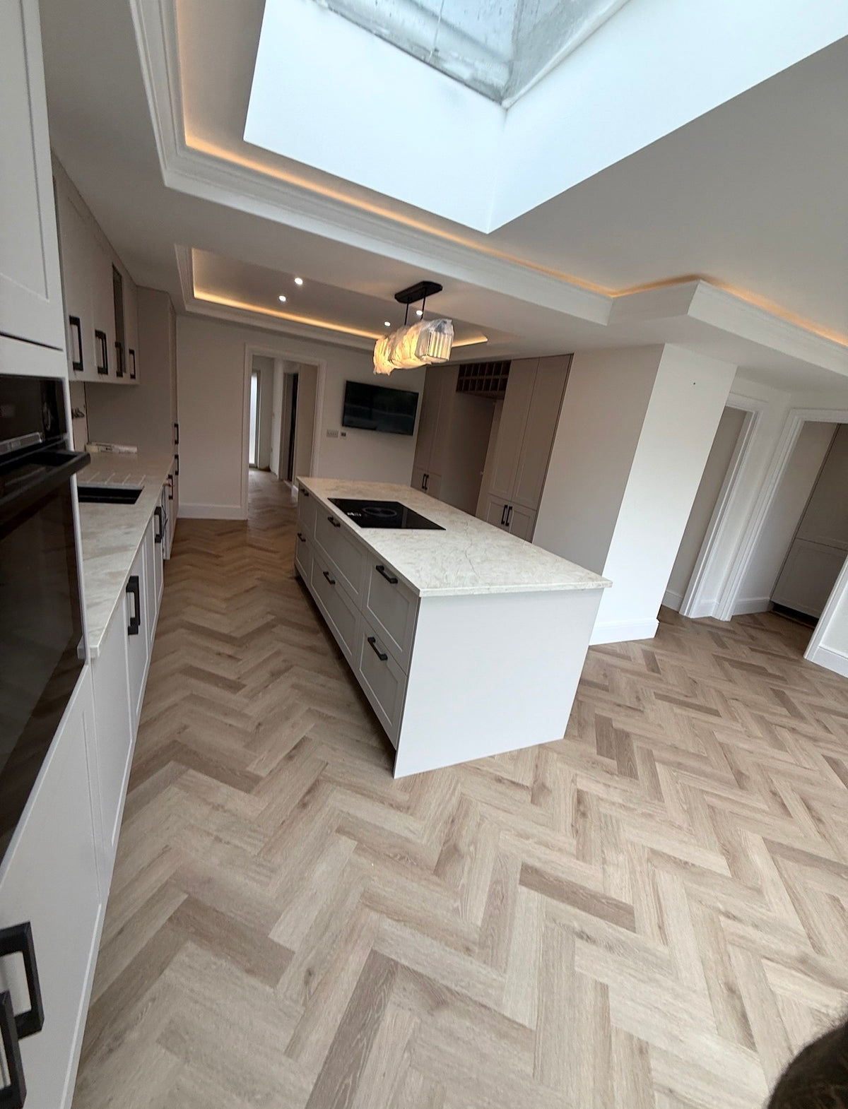 A modern kitchen interior with light beige herringbone wood flooring, white cabinetry, a central island with a light stone countertop, recessed lighting, and a large skylight.