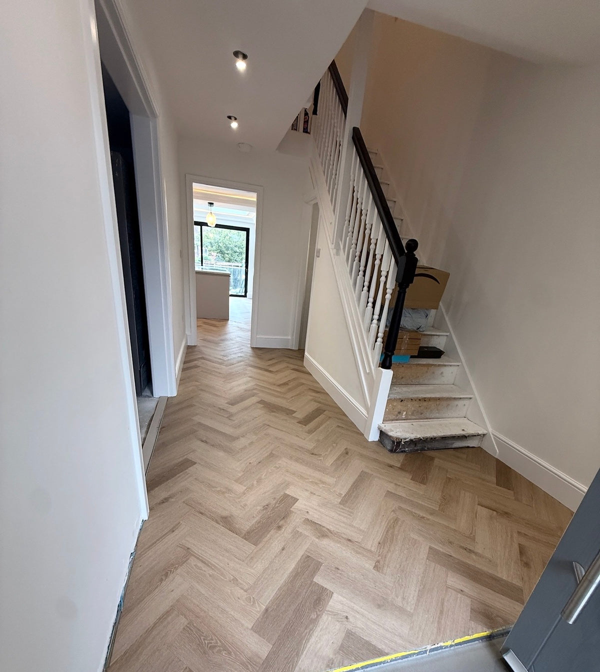 A modern interior hallway with light oak herringbone wood flooring, featuring a staircase with white steps and a black handrail on the right, and a doorway leading to another room in the distance.