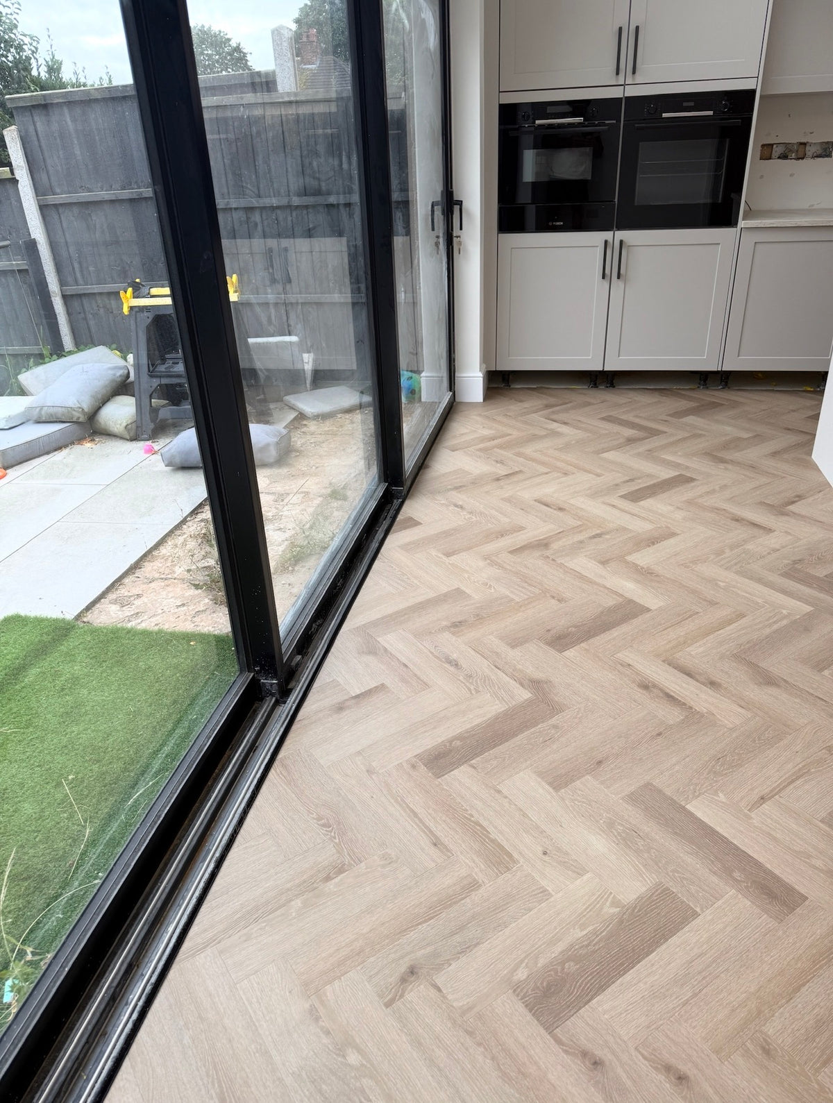 Light oak herringbone wood flooring installed in a modern kitchen and patio area, with large black-framed sliding glass doors showing a backyard.