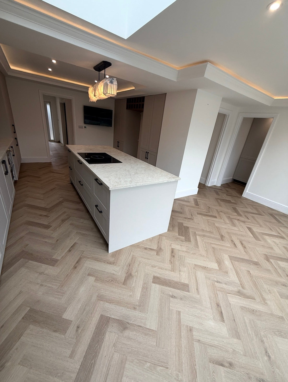 A modern kitchen interior with a large white island featuring a light beige marble-look countertop and a black cooktop. The room has white walls, recessed lighting, and a prominent herringbone wood floor in a light oak tone.