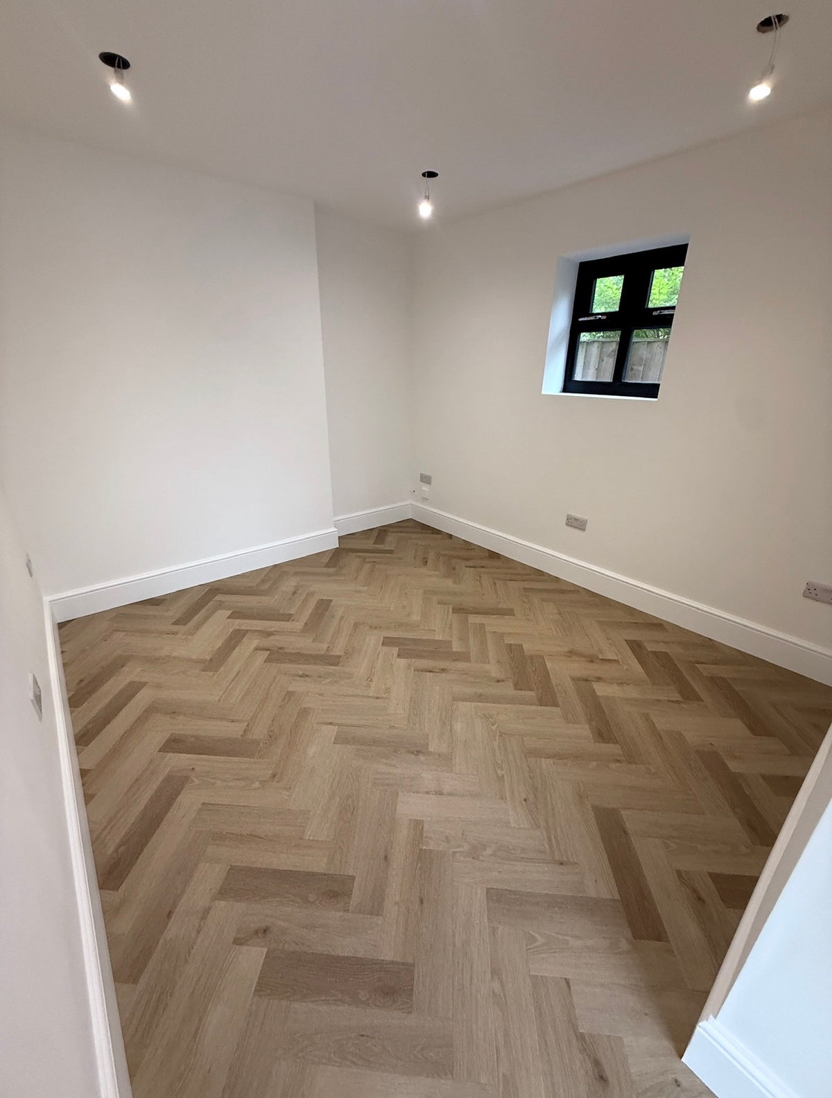 A room with light oak herringbone parquet flooring, white walls, and a small black-framed window.