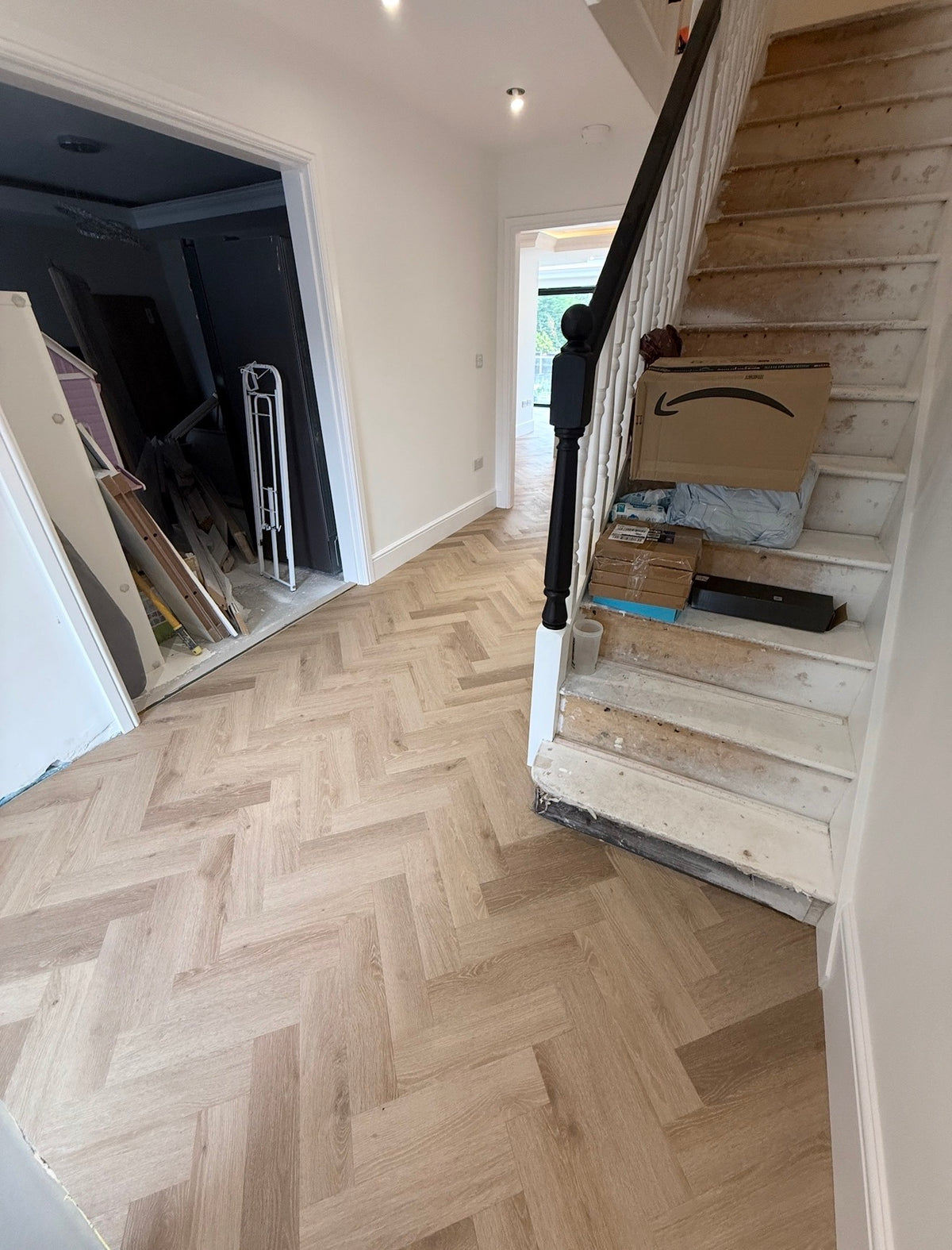 Light oak herringbone engineered wood flooring installed in a home stairwell, with a wooden staircase and black banister on the right and an open doorway on the left.