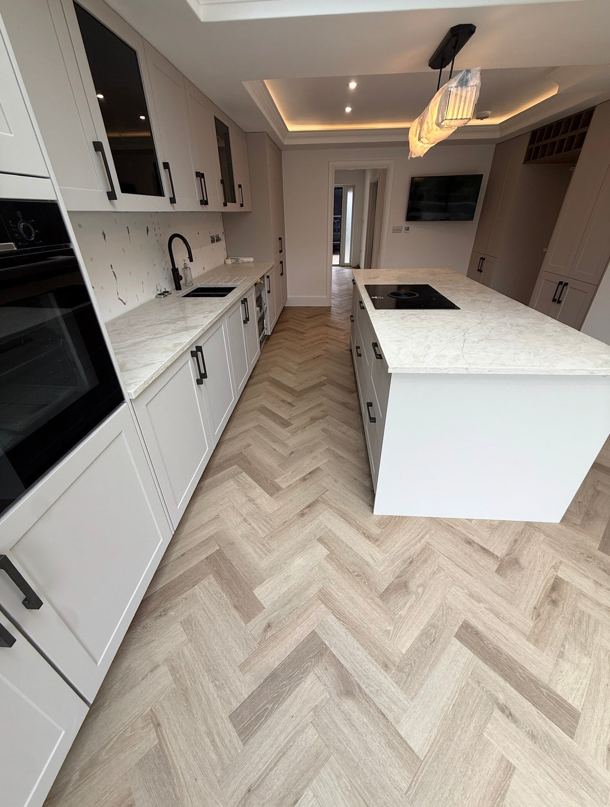 A modern kitchen interior with white cabinetry, marble countertops, a central island, and light wood herringbone flooring.