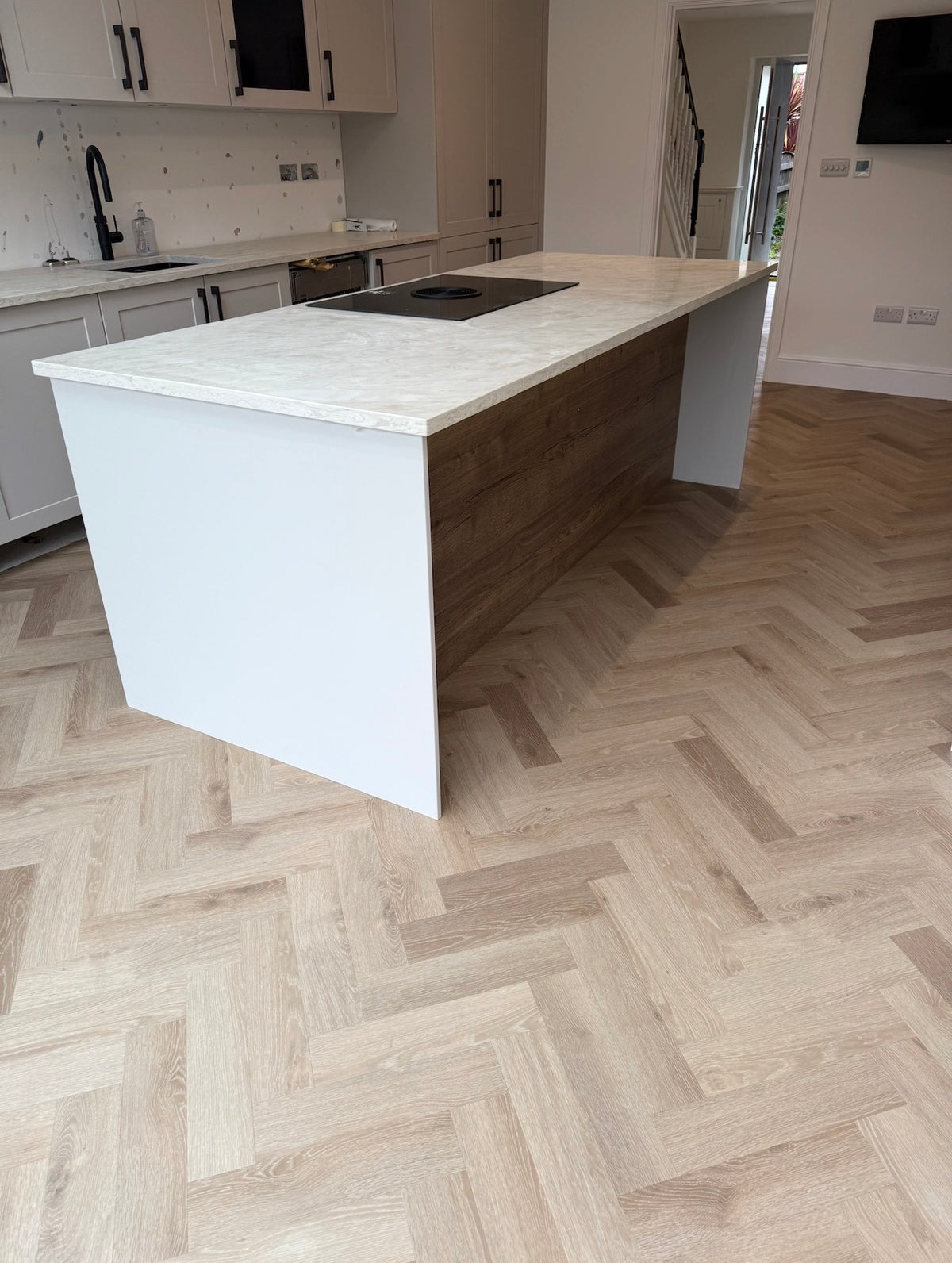 A modern kitchen island with a white front and a light wood side panel, featuring a white marble-look countertop and a black built-in cooktop, set on a light herringbone wood floor.